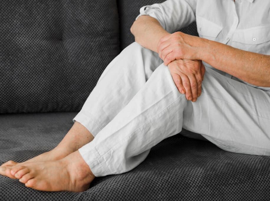 An elderly person sitting, hands clasped over one knee, possibly indicating pain