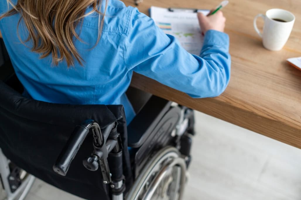 Person in a wheelchair writing on a document at a table with a cup of coffee in the background